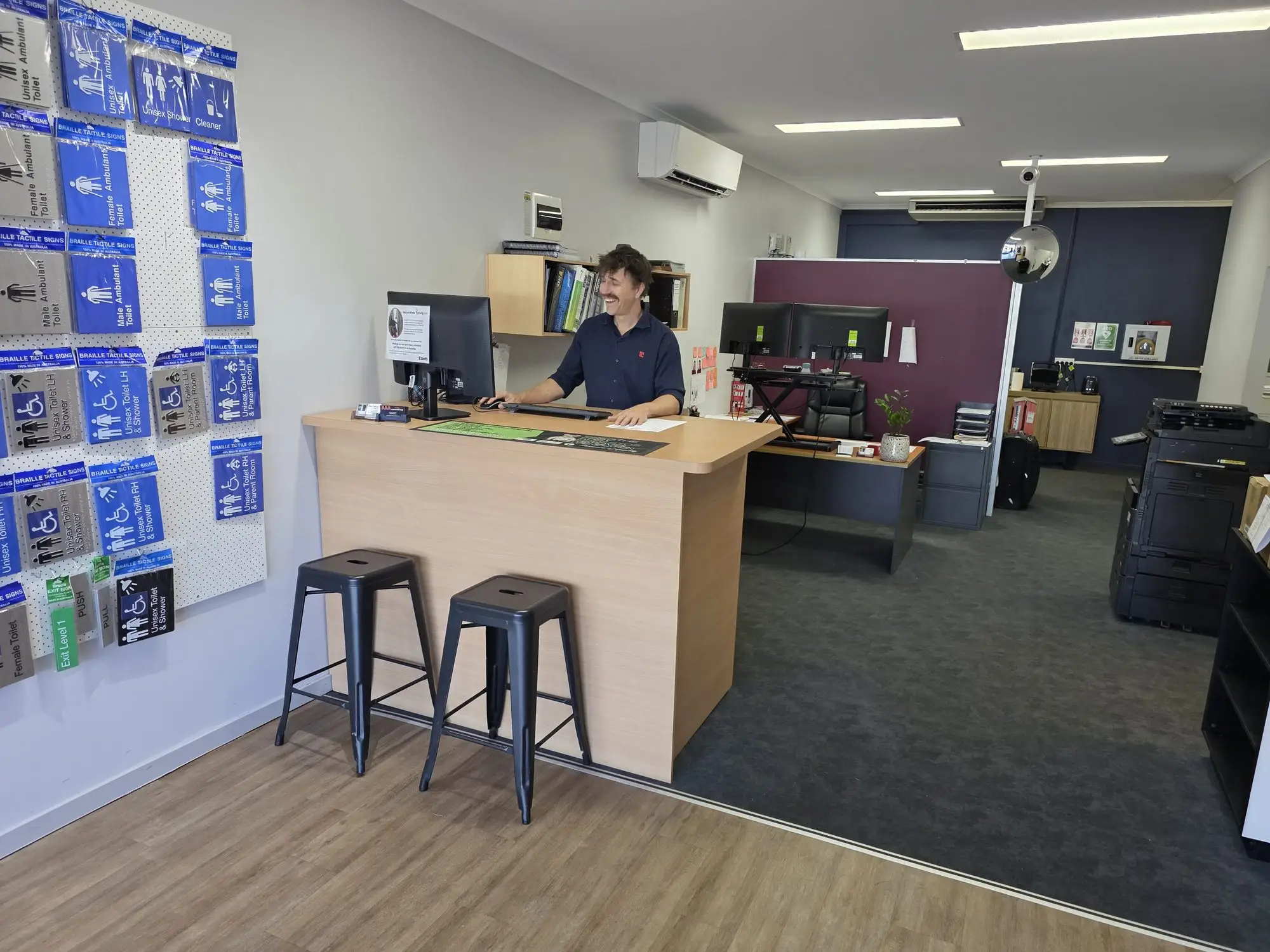 A Bretts PHD team member at the front counter, beside a wall of braille and tactile washroom signs.
